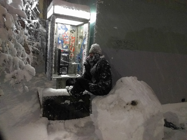A person in snowy clothing sits on a giant snowball and talks into public Futel telephone.